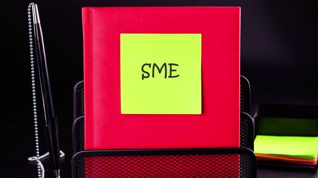 A bright green sticker with text animation SME,SMEs (or small and medium enterprises) is placed on a red notebook, which emphasizes encouragement for initiative in a professional work environment