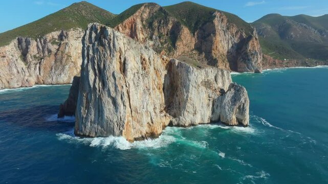 Pan di Zucchero, the towering sea stack off Masua, Sardinia with sharp limestone walls rising from the vibrant waters, backed by steep coastal cliffs and green Mediterranean hills, dramatic drone shot
