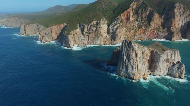 Aerial pull outfootage of Pan di Zucchero, the iconic sea stack near Masua, Sardinia. Towering limestone cliffs rise from deep blue waters, backed by green hills under clear Mediterranean skies.
