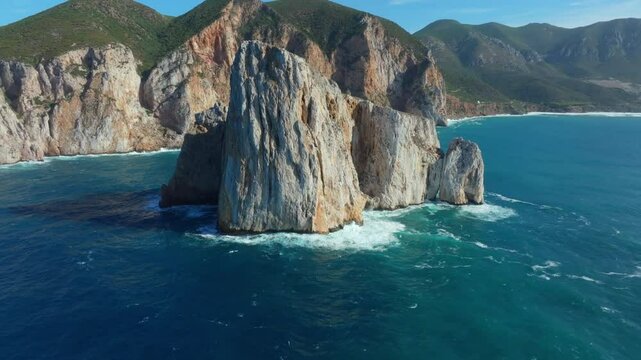 Slow Drone footage captures Pan di Zucchero rising from turquoise waters near Masua, Sardinia, Sheer rock faces meet rolling green hills and the vibrant Mediterranean Sea in stunning coastal harmony.