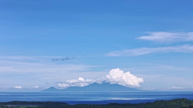 A sweeping panoramic shot captures Camiguin Island shrouded in morning clouds as seen from the mainland of Mindanao, Philippines, with clear blue skies and the ocean below.