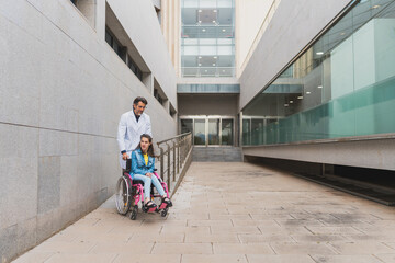 Young woman with cerebral palsy pushed by doctor outside building - Medical professional assists female patient in wheelchair on accessible ramp. Concept of cerebral palsy.