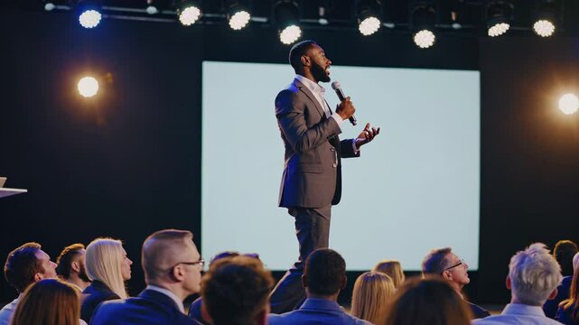 Confident black businessman walks and speaks on stage at a high-level business conference, engaging a large audience with his inspiring, dynamic speech. Concept transformative influence.
