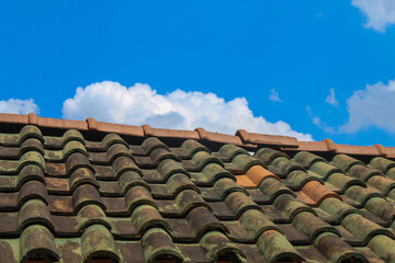 A close-up view of a traditional tiled roof under a clear blue sky with scattered clouds. The moss-covered tiles add texture and contrast to the vibrant background.