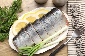 Pieces of delicious herring with lemon slices, green onions and dill on wooden table, flat lay