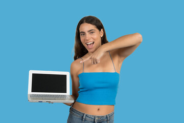 Young woman pointing at laptop with blank screen on blue background