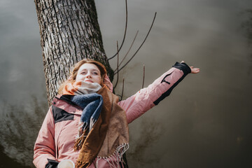 A woman in a cozy jacket and scarf lies contentedly by a tree next to a calm water body, expressing a sense of relaxation and connection with nature in an outdoor leisure moment.