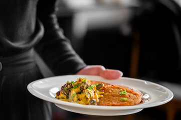 A close-up of a server holding a stylish plate featuring golden-brown cutlets paired with creamy pasta and garnished with fresh herbs, showcasing an elegant dining experience