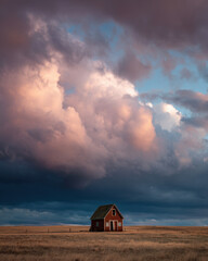 A small barn house rests in a vast field as vibrant clouds loom overhead during sunset, creating a serene and tranquil atmosphere