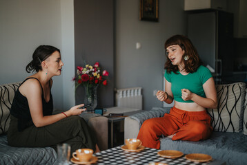 Two young women engage in a cheerful conversation, sitting comfortably in a cozy living room with a...