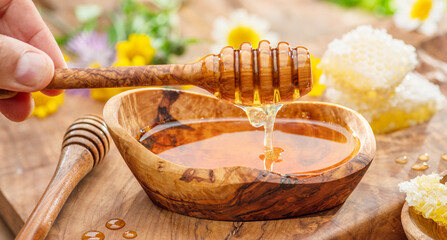 Honey dripping from wooden spoon into wooden bowl with honey, spring flowers are on wooden table...