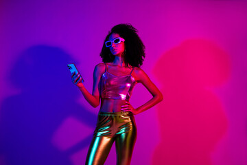 Fashionable woman with a stylish metallic outfit and sunglasses holding a smartphone, illuminated by vibrant neon purple and pink lights