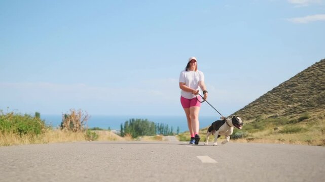 A young curvy woman in sportswear walks along the road with a pit bull pulling the leash, tongue out from the heat. Green hills surround them, the sea in the distance, with the sun lighting their way.