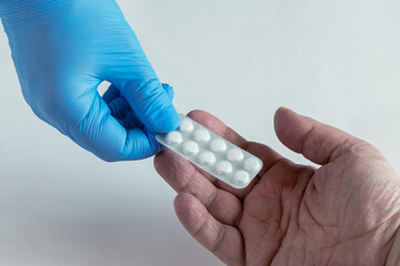 The hands of a doctor or pharmacist in blue medical gloves extending a blister pack of white pills...