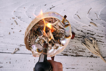 Burning sweetgrass (Hierochloe odorata) and white sage (Salvia apiana) in an iridescent abalone shell on an old white wooden table. Overhead view.