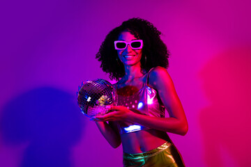 Naklejka premium Portrait of a vibrant young woman in metallic attire holding a disco ball against neon lights, evoking night club energy and retro party
