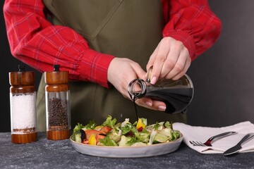 Woman pouring balsamic vinegar onto tasty salad at dark textured table, closeup