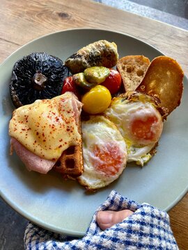 Close-up photograph of a traditional cooked breakfast served on a ceramic plate, featuring fried eggs, sausages, toast, mushrooms, tomatoes and other classic breakfast foods. 