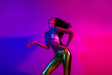 Young stylish woman in a fashionable outfit dancing under vibrant neon lights with glowing purple and pink tones on the background.