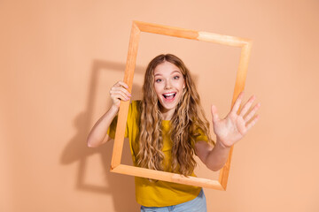 Joyful young woman holding wooden frame smiling in casual clothes against beige backdrop expressing fun and happiness