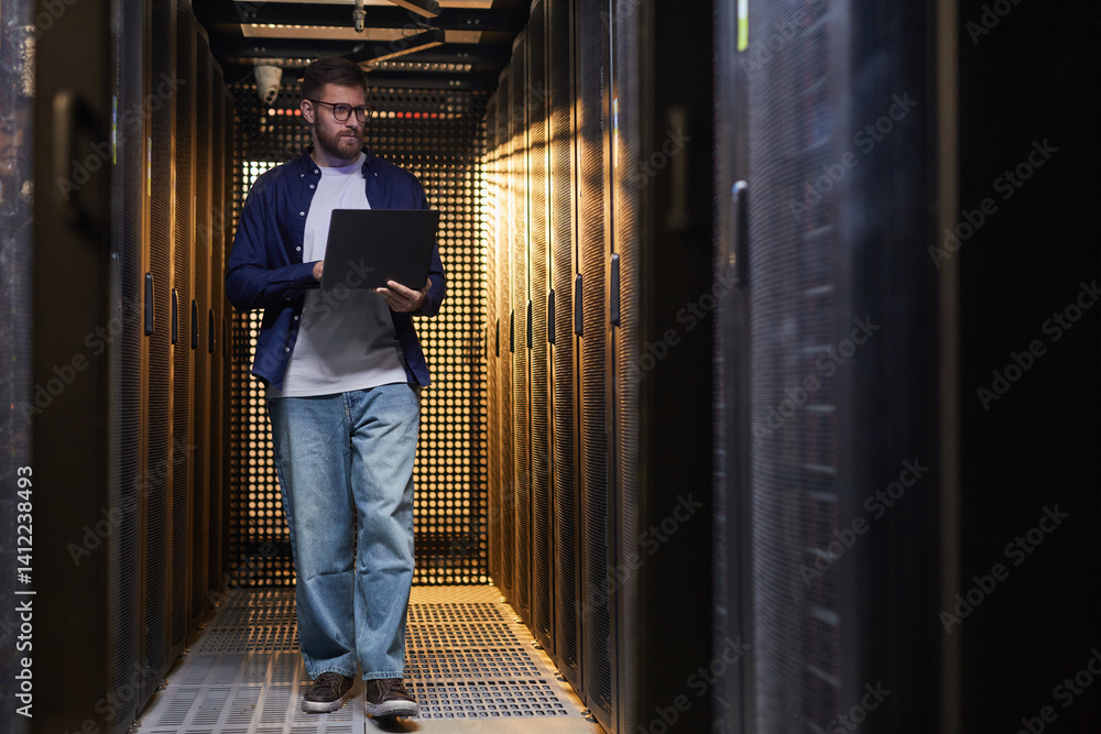 Poster technician standing in server aisle while monitoring data systems on laptop. surrounded by server ra