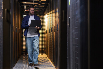 Technician standing in server aisle while monitoring data systems on laptop. Surrounded by server...