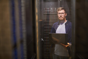 Portrait of technician standing in server room holding laptop and managing server equipment....