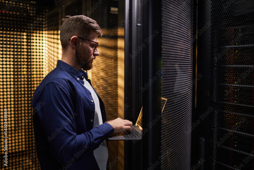 Poster profile of a man engaged in work on laptop while in modern server room with rows of servers. focused