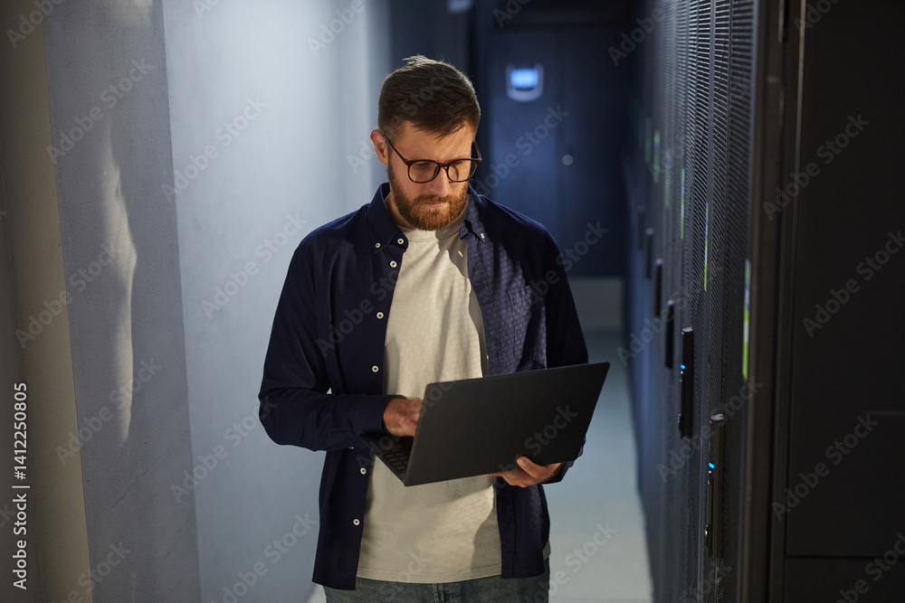 Poster man with beard and glasses standing in a dark corridor of a data center, working on laptop while ins