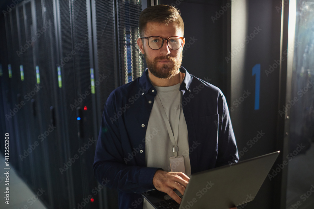Poster portrait of man with beard wearing glasses and holding laptop in a server room with rows of server r