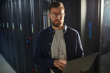 Portrait of man with beard wearing glasses and holding laptop in a server room with rows of server...