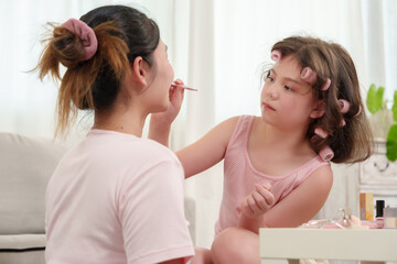 Fototapeta premium Caucasian girl using lipstick pencil on lips of smiling Asian woman while sitting closely together indoors showing playful learning moment bonding love trust and joy between mother and daughter