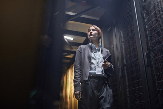 Woman standing in data center, checking servers and holding a device, with focused expression and professional attire, surrounded by technology and cables