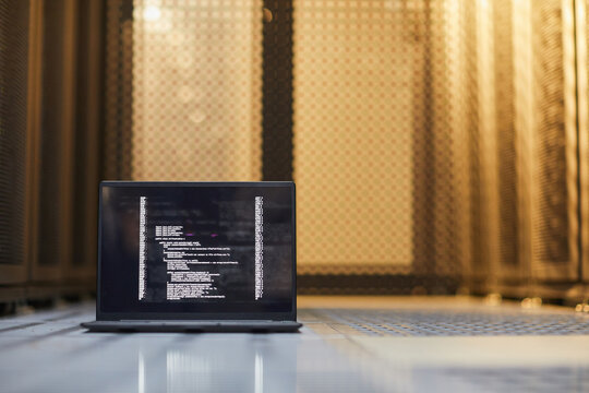 Open laptop displaying lines of code against backdrop of server racks in dim lighting. Scene represents data processing, programming, and technology infrastructure maintenance