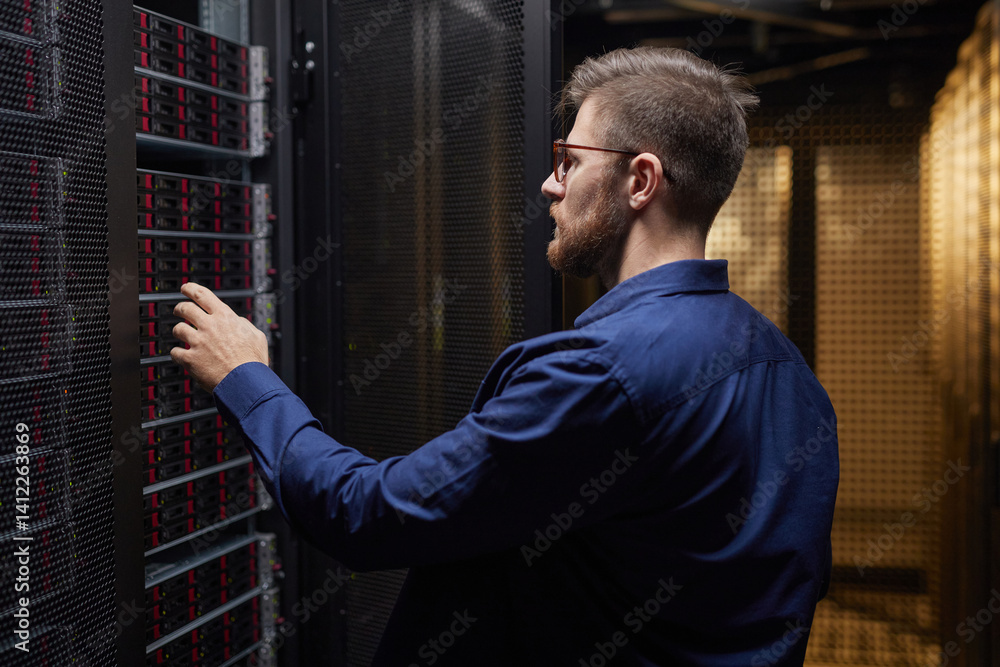 Poster technician adjusting server hardware in modern data center facility with several racks visible. pers