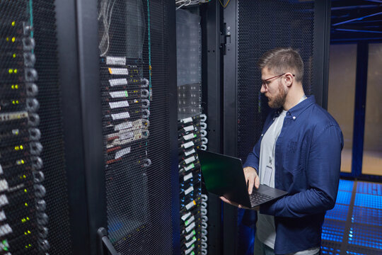 Technician standing in server room checking connections and status of servers using laptop while surrounded by multiple racks of equipment and cables