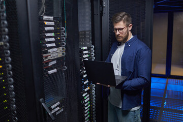 Male IT technician standing in modern server room holding laptop, checking network equipment and...