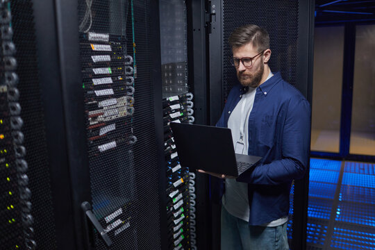 Male IT technician standing in modern server room holding laptop, checking network equipment and ensuring proper functionality. High-tech environment featuring server racks and data cables