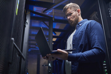 Technologist wearing glasses holding laptop standing in a data center, engaging with server...