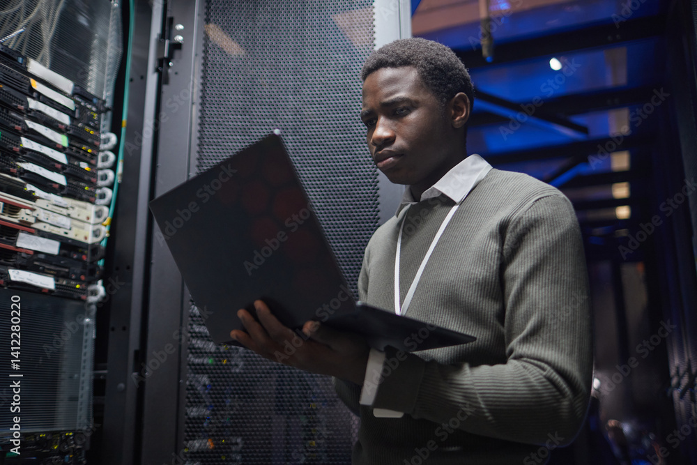 Poster african american technician working with a laptop inside data center server room, analyzing server p
