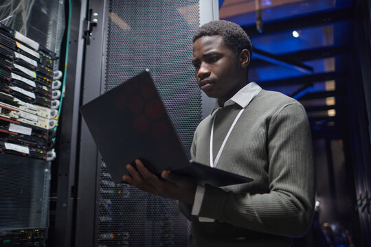 African American technician working with a laptop inside data center server room, analyzing server performance, monitoring systems and infrastructure, ensuring network stability and connectivity