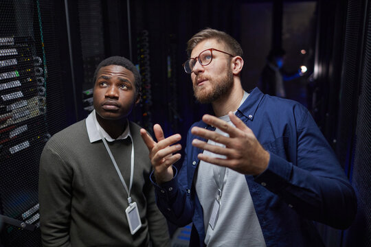 Two technicians in data center, one African American, standing near server racks while discussing maintenance process, one person gesturing with hand, showing attentiveness - Powered by Adobe