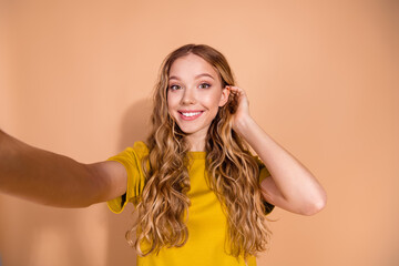 Playful young woman with wavy blonde hair taking a cheerful selfie against a beige background