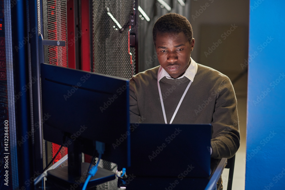 Poster focused black person working with a laptop in a data center surrounded by server racks and cables in