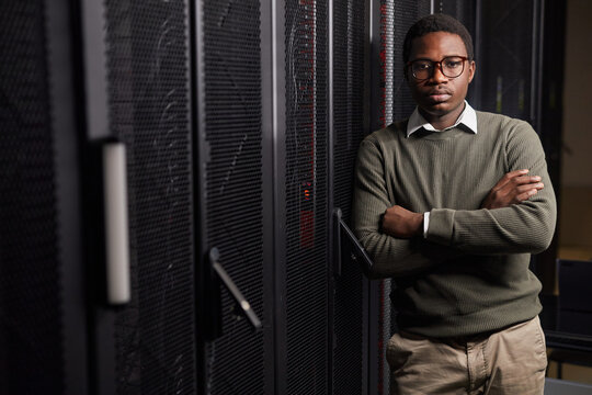Portrait of confident young African American male standing in a data center, crossing arms and looking at camera, wearing glasses and casual sweater beside server racks with calm expression