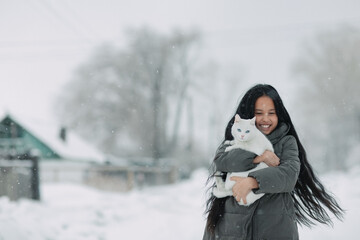 A girl with a white cat stands joyfully under the snow