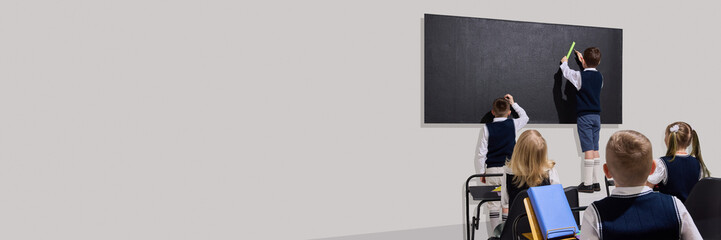 Kids standing at blackboard, solving problem using ruler while classmates watch attentively from...