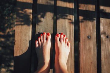 a person's feet with red nail polish resting on wooden planks in the sunlight, creating shadows.