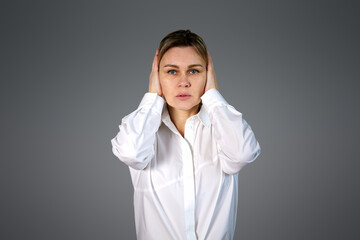 A blonde girl covered her ears with her hands, a woman in a white shirt on a gray background. Woman on Dark Background with Copy Space in White Office Shirt, Business, Business Style, Concept