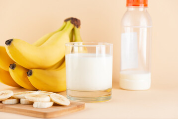 Banana milk in a glass and bottle with bananas slices in a wooden board on a beige background....